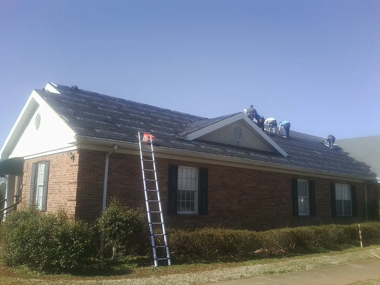 Crew tearing shingles off a brick ranch roof with a ladder set — Emergency Roof Repair work by our crew in Manhasset
