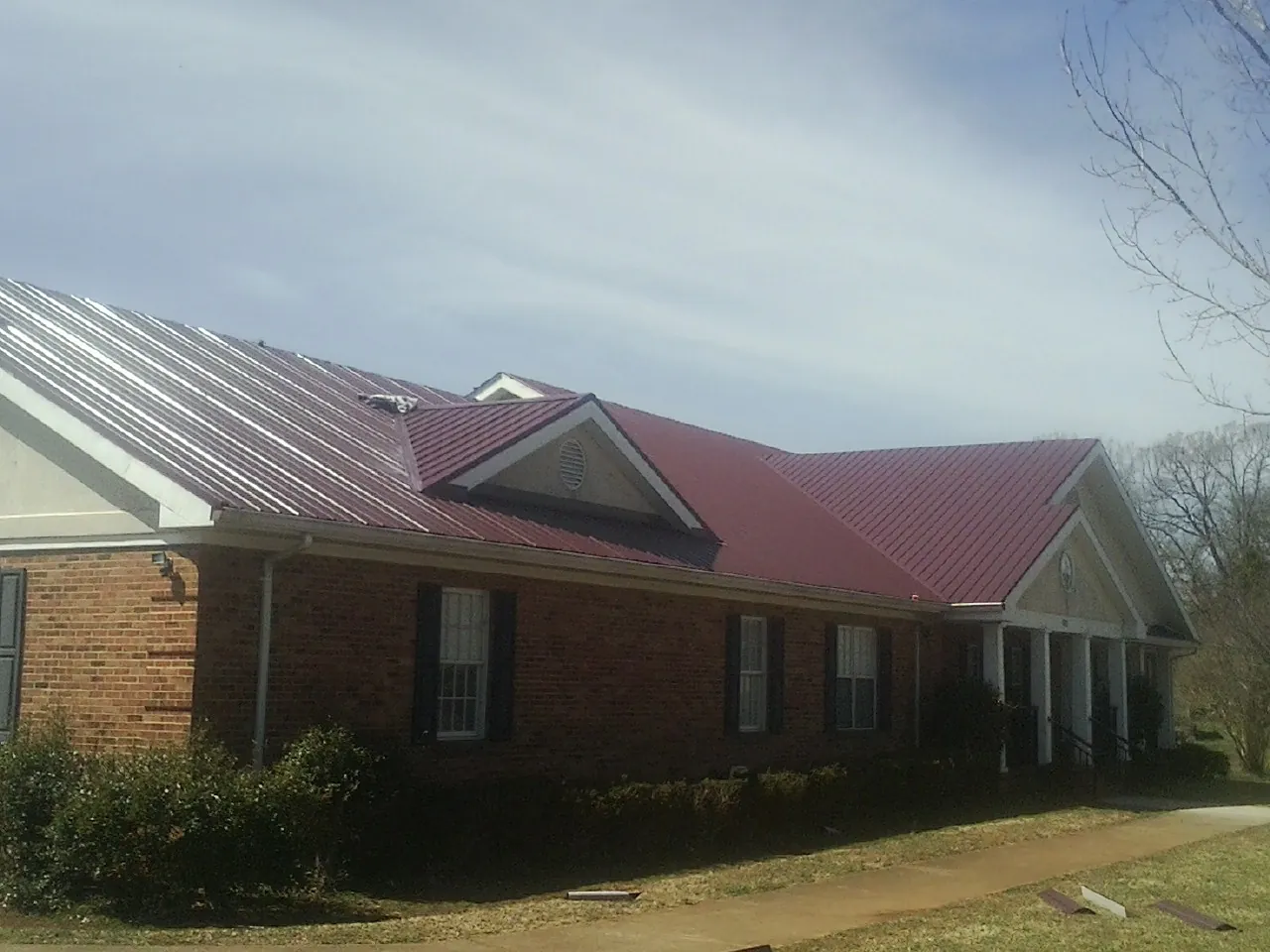 Red metal roof with gabled entries on a long brick building, from a Hail Damage Roof Repair job in Manhasset