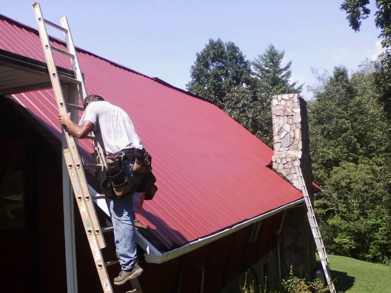 Roofer on a ladder beside a red metal roof and stone chimney — Soffit & Fascia Repair by Roofing Brothers in Manhasset